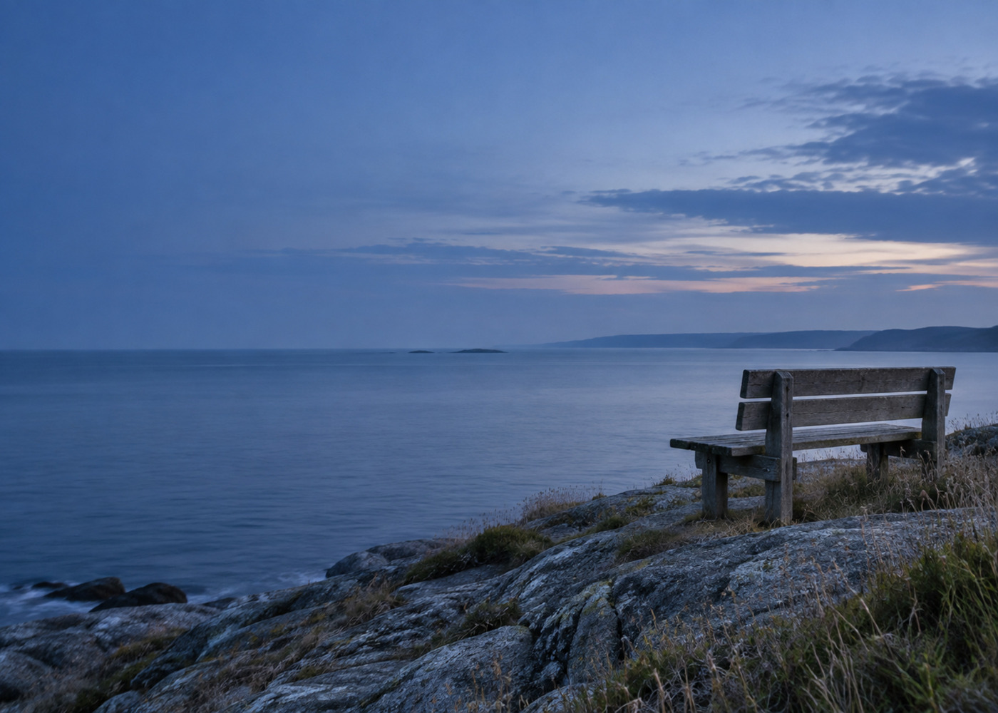 An empty weathered bench on a rocky headland looking out over calm water at dusk